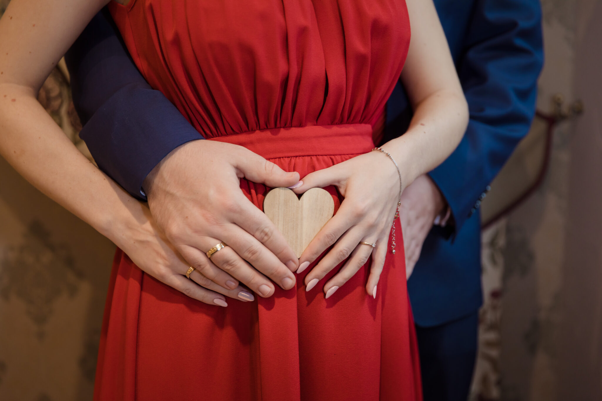 sensownipl duet fotograficzny z Krakowa. Fotografia z sesji ciążowej. Bliski kadr na ciążowy brzuszek i dłonie rodziców wspólnie trzymających drewniane serduszko.